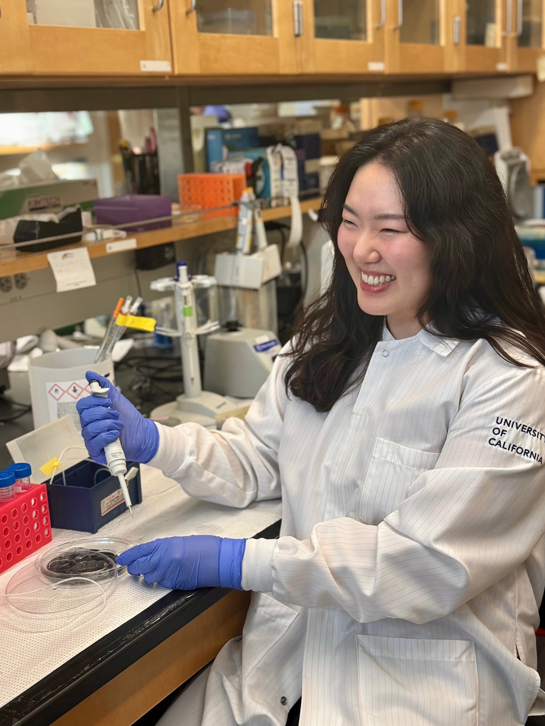 A woman wearing a lab coat and gloves smiles 