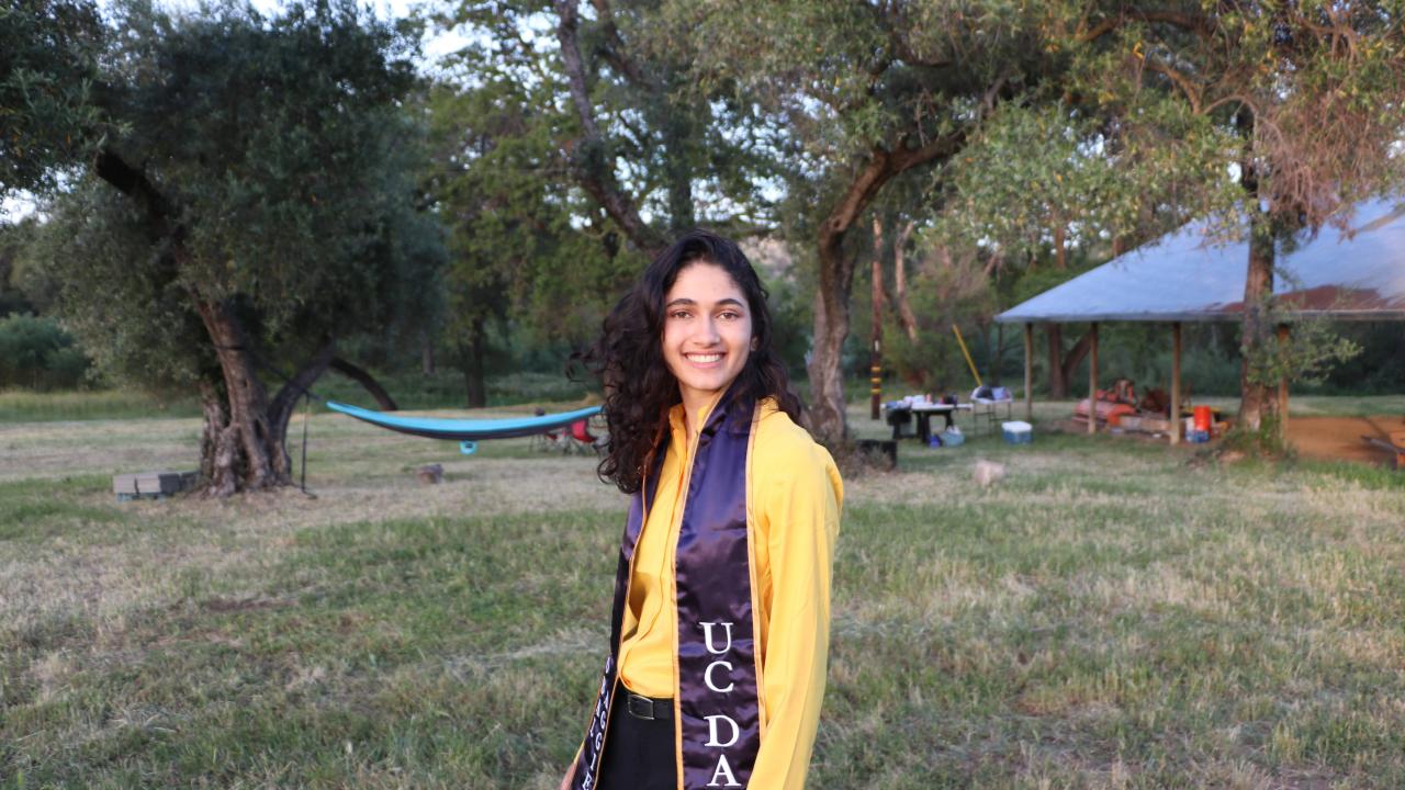 A woman wearing a UC Davis stole smiles in a grassy field 