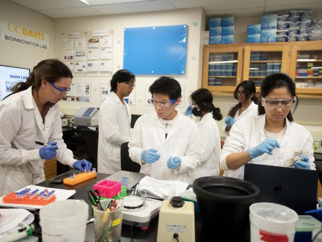 Five people in white lab coats and goggles working at a crowded lab bench, wearing blue gloves.