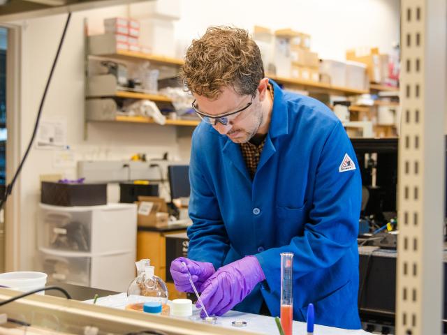 Lab worker wearing safety glasses and purple gloves pipetting at a cluttered bench.