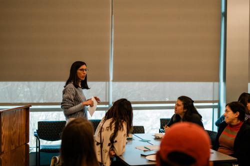 A woman holding a paper stands at the front of a meeting room addressing other women seated at a table