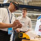 One person wears augmented reality goggles while another stands close by near a poster and interactive activity